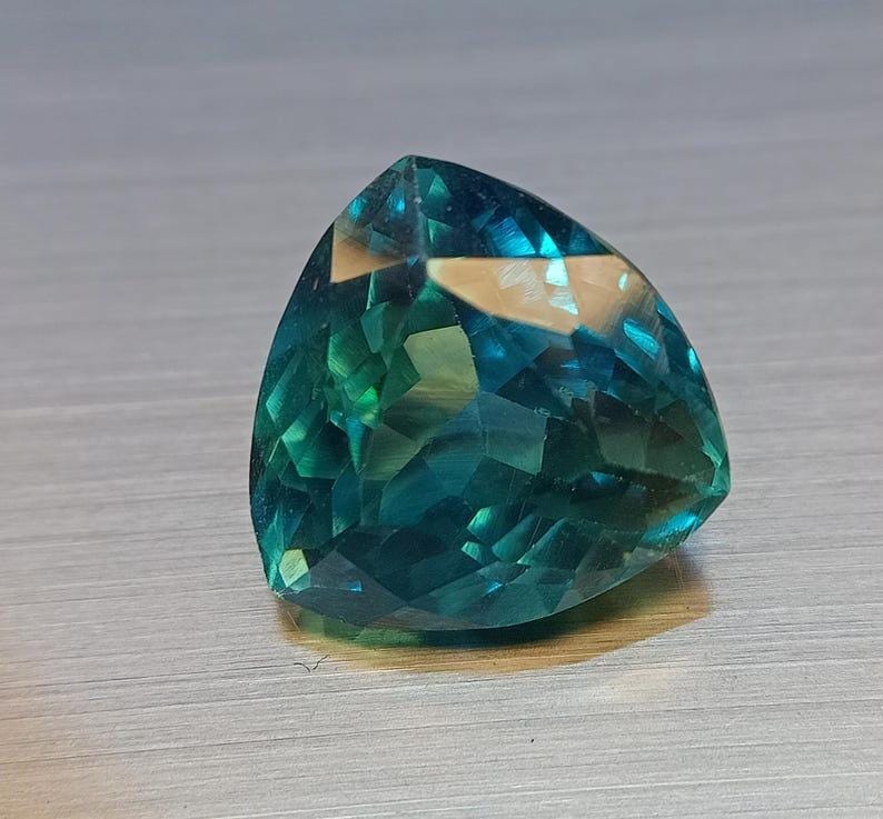 May include: A triangular-cut gemstone with facets, displaying shades of teal and green. The gem has a polished surface and reflects light, creating a sparkling effect. The gemstone is set against a neutral background.