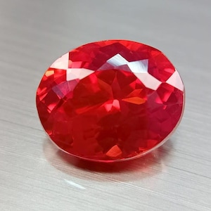 Può includere: Una gemma rossa sfaccettata, di forma ovale. La gemma ha un taglio brillante, che riflette la luce con numerose sfaccettature. Il colore è un rosso vibrante e intenso, con riflessi bianchi. La gemma è posta su una superficie grigio chiaro.