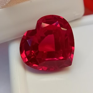 Puede incluir: Una gema facetada en forma de corazón, de un tono rojo vibrante. El corte de la gema revela múltiples facetas, creando un efecto brillante. La gema se encuentra sobre una superficie blanca.
