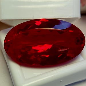 May include: A vibrant, oval-cut gemstone with a deep, saturated red color and multifaceted surface that reflects light. The stone is positioned resting within a white, rectangular display box with the lid partially open behind the gem.