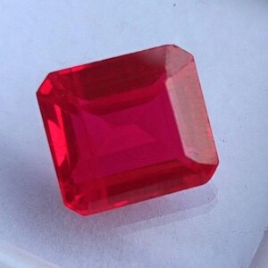 Può includere: Una gemma rossa vibrante, a taglio quadrato. La gemma è di un colore rosso intenso e ricco e sembra trasparente. La forma è un taglio smeraldo modificato, con una faccia rettangolare e angoli smussati.
