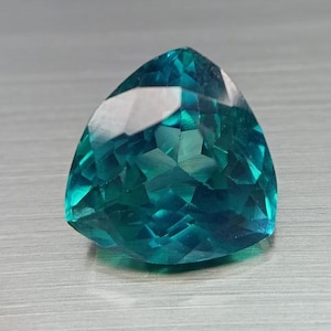 May include: A faceted, triangular gemstone with a vibrant teal color. The gem has a polished surface with multiple facets that reflect light, creating a sparkling effect. The gemstone is set against a neutral, textured background.