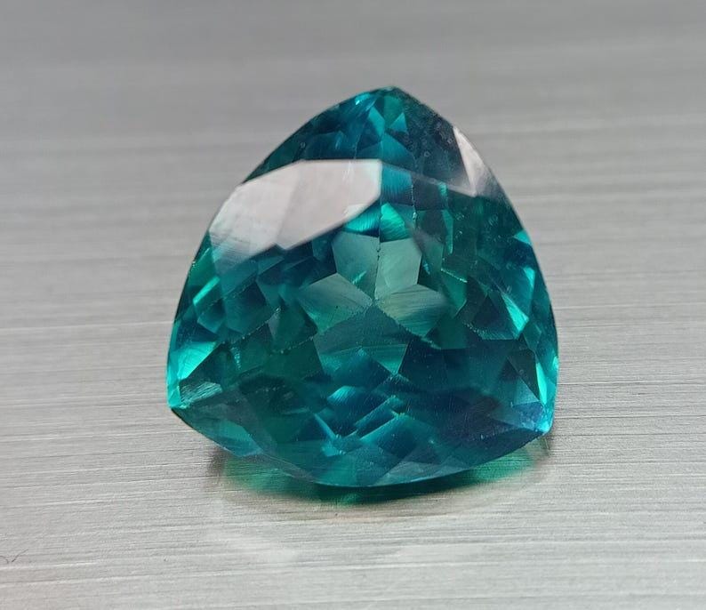 May include: A faceted, triangular gemstone with a vibrant teal color. The gem has a polished surface with multiple facets that reflect light, creating a sparkling effect. The gemstone is set against a neutral, textured background.