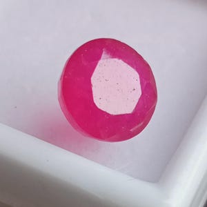 Puede incluir: Una gema facetada, redonda, de color rosa intenso. La gema tiene un centro claro, de forma hexagonal irregular. La gema está colocada sobre un fondo blanco.