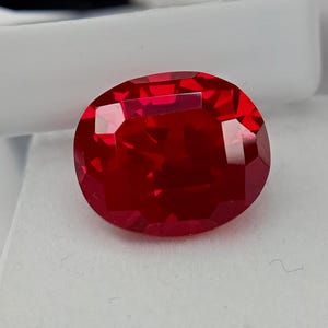 Könnte beinhalten: Ein leuchtender, oval geschliffener Edelstein in einer tiefen, gesättigten roten Farbe. Der Stein weist zahlreiche geometrische Facetten auf, die das Licht reflektieren und eine brillante, funkelnde Optik auf der polierten, durchscheinenden Oberfläche erzeugen.