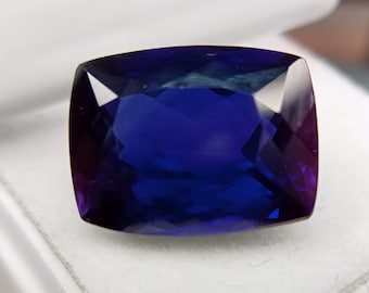 Può includere: Una gemma sfaccettata, taglio a cuscino, con un centro blu intenso e bordi rosso-viola. La gemma è posta su uno sfondo bianco, che ne mette in risalto la chiarezza e la brillantezza. Il taglio della pietra crea molteplici superfici riflettenti.