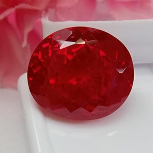 Può includere: Una gemma dal taglio ovale di colore rosso intenso con una superficie sfaccettata che riflette la luce. La pietra è posizionata su una superficie bianca liscia, mettendo in mostra la sua brillante chiarezza e i precisi tagli geometrici.