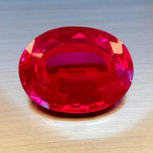 May include: A faceted, oval-shaped, red gemstone. The gem has a high degree of clarity and brilliance, reflecting light across its many facets. The gemstone is a deep, rich red colour, with a polished surface.