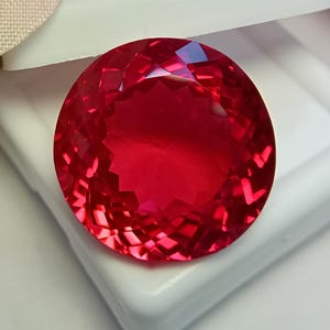 May include: A round, faceted gemstone with a vibrant, deep red color. The stone features a complex, geometric cut that creates numerous light-reflecting surfaces across its circular face, showcasing a brilliant, saturated crimson hue throughout.