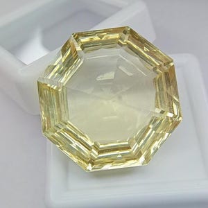 May include: A faceted, octagonal gemstone with a pale yellow hue. The gem is set in a white plastic container. The facets create a play of light and shadow, enhancing the gem's brilliance. The gem's shape is symmetrical and geometric.