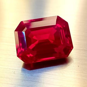 May include: A vibrant, rectangular-cut ruby gemstone. The gem is a deep red hue, with precisely cut facets that catch the light. The gemstone is displayed on a neutral, metallic surface.
