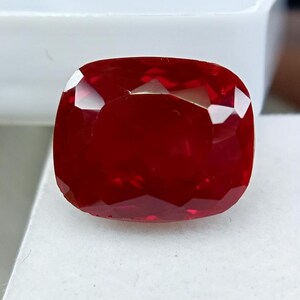 Peut inclure: Une pierre précieuse de type rubis, taillée en coussin, avec de multiples facettes. La gemme est d'un rouge profond et saturé. La pierre est posée sur un fond blanc.