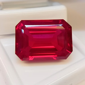 Könnte beinhalten: Ein facettierter, rechteckig geschliffener Rubin-Edelstein. Der Edelstein hat eine leuchtend rote Farbe und eine hohe Klarheit. Die Facetten reflektieren das Licht und erzeugen ein brillantes Aussehen. Der Rubin liegt auf einem weißen Hintergrund.