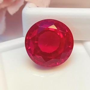 May include: A faceted, round, deep red gemstone, possibly a ruby, is the focal point of this image. The gem is set against a white background, with a hint of a pink flower in the upper left corner. The gem's facets reflect light, creating a brilliant display.