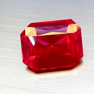 Puede incluir: Una gema roja vibrante, tallada en forma rectangular y facetada. La gema tiene un alto grado de claridad, con bordes afilados y una superficie pulida. La gema se presenta sobre un fondo claro, resaltando su brillo.