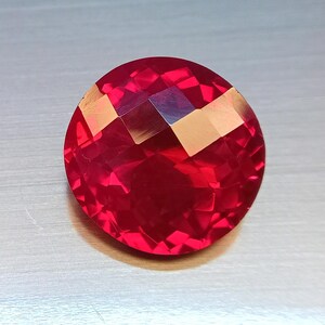 May include: A round, faceted gemstone with a vibrant, deep red color. The stone features a complex geometric cut with numerous triangular and quadrilateral facets that create a high level of light reflection and internal brilliance across its surface.