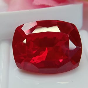 May include: A faceted, cushion-cut, deep red gemstone. The gem is displayed on a white surface, with a soft pink background. The gemstone's facets reflect light, creating a brilliant display of color and sparkle.