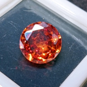 May include: A faceted, round gemstone with a fiery orange-red hue. The gem is set against a dark background within a white-edged container, showcasing its brilliance and clarity. The cut of the stone creates multiple facets that reflect light.