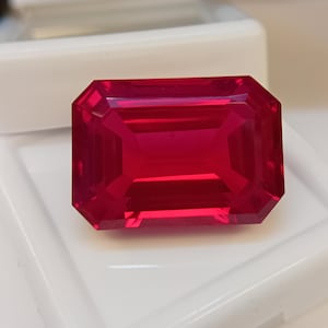 May include: A faceted, rectangular ruby gemstone with a deep red hue. The gem is set against a white background, showcasing its clarity and brilliance. The cut of the stone highlights its internal structure and light refraction.