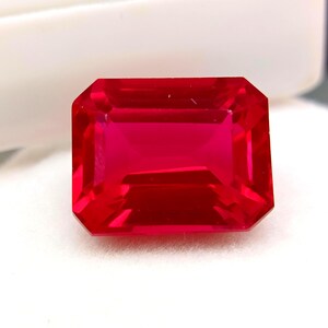 Può includere: Una vibrante gemma rosso rubino, taglio rettangolare. La gemma ha una superficie sfaccettata che riflette la luce, mostrando la sua chiarezza e brillantezza. La gemma è posta su uno sfondo bianco, evidenziando il suo colore e la sua forma.