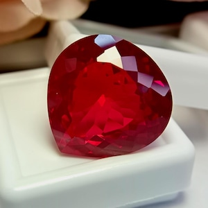 Könnte beinhalten: Ein leuchtend roter, herzförmiger Edelstein mit einer facettierten Oberfläche, die das Licht reflektiert. Der Stein zeigt einen tiefen, gesättigten Karmesinton mit scharfen, geometrischen Schliffen auf der Oberseite, die für Glanz sorgen.