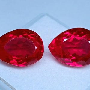 Puede incluir: Dos gemas facetadas, rojas brillantes, en forma de pera. Las gemas se muestran una al lado de la otra en un recipiente cuadrado blanco. Las piedras tienen un alto brillo y probablemente sean rubíes, con un corte brillante.