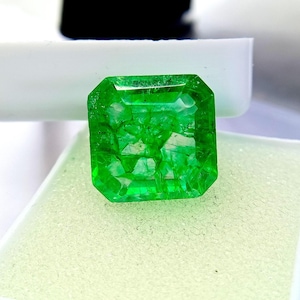 Könnte beinhalten: Ein lebendiger, smaragdgeschliffener grüner Edelstein. Der Edelstein hat eine leuchtende, satte grüne Farbe mit sichtbaren inneren Einschlüssen. Er ist vor einem weißen und hellgrünen Hintergrund platziert, der seine Klarheit und Brillanz hervorhebt. Der Edelstein hat eine quadratische Form mit abgeschrägten Kanten.