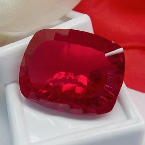 May include: A large, cushion-cut ruby gemstone with a deep red hue. The gem is displayed in a white tray, with light reflecting off its many facets. The background features white and red fabric. The ruby is a vibrant, eye-catching piece.
