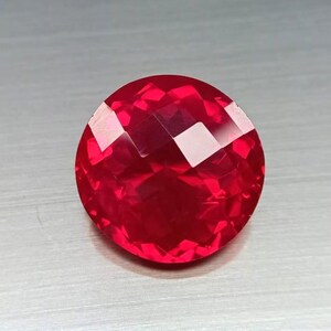 May include: A faceted, round, red gemstone. The gem has a brilliant, reflective surface with multiple facets that catch the light. The gemstone is a deep, rich red color, and appears to be polished.