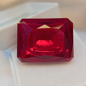 May include: A faceted, rectangular-cut ruby gemstone. The gem is a deep, saturated red color, with sharp, clean facets that reflect light. The ruby is set against a white background, highlighting its brilliance and clarity. The gemstone is a symbol of passion and luxury.