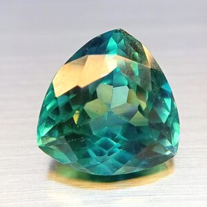 May include: A faceted, triangular gemstone with a vibrant teal and green color palette. The gem's surface reflects light, showcasing its intricate cuts and angles. The gemstone is a striking example of fine jewelry.