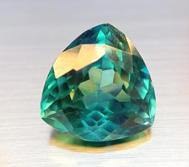 May include: A faceted, triangular gemstone with a vibrant teal and green color palette. The gem's surface reflects light, showcasing its intricate cuts and angles. The gemstone is a striking example of fine jewelry.