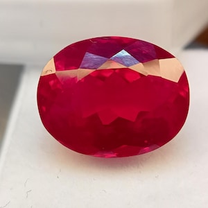 Può includere: Una gemma di rubino sfaccettata a forma ovale. La gemma è di un colore rosso intenso e saturo, con molteplici sfaccettature che riflettono la luce. La gemma è posta su uno sfondo bianco, evidenziando la sua brillantezza e il suo colore.