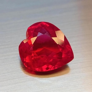 Könnte beinhalten: Ein facettierter, herzförmiger, roter Edelstein. Der Stein hat mehrere reflektierende Oberflächen, die das Licht einfangen und einen funkelnden Effekt erzeugen. Die Herzform ist klar definiert, mit einem glatten, polierten Aussehen. Der Edelstein liegt auf einer hellen Oberfläche.