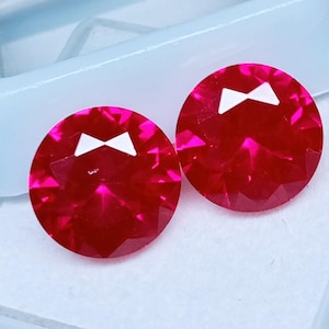 May include: Two round, faceted, bright red gemstones with a white bow-tie shaped design. The gems are set against a white background, suggesting a jewelry or craft supply theme.