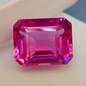 May include: A vivid, faceted, emerald-cut gemstone in a deep pink colour. The gem has a rectangular shape with beveled edges, reflecting light and showcasing its internal structure. The gem is set against a white background.
