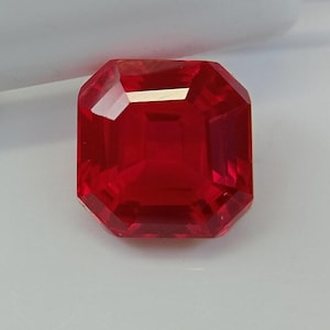 Könnte beinhalten: Ein facettierter, rechteckig geschliffener, roter Edelstein. Der Edelstein hat eine leuchtende, tiefrote Farbe und ist vor einem weißen Hintergrund platziert. Der Schliff des Edelsteins erzeugt mehrere Facetten, die das Licht reflektieren.