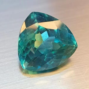 May include: A faceted, triangular gemstone with a vibrant teal and turquoise color. The gem's surface reflects light, showcasing its many facets and internal structure. The gemstone is set against a neutral background, highlighting its brilliance.
