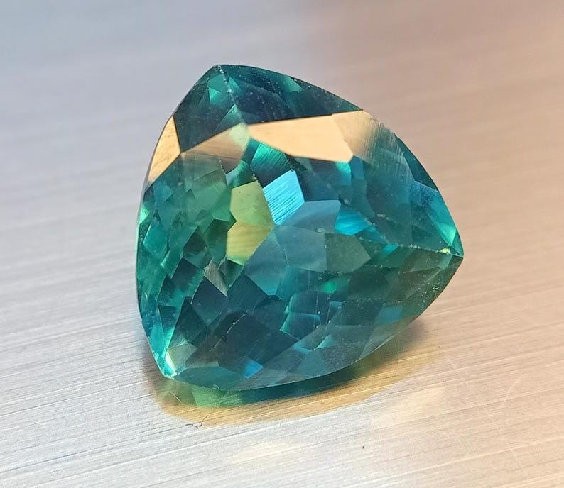 May include: A faceted, triangular gemstone with a vibrant teal and turquoise color. The gem's surface reflects light, showcasing its many facets and internal structure. The gemstone is set against a neutral background, highlighting its brilliance.