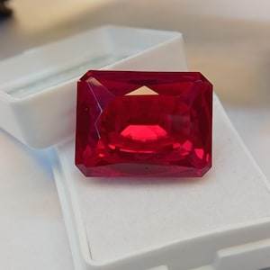 Könnte beinhalten: Ein großer, rechteckiger, facettierter Rubin-Edelstein mit tiefroter Farbe. Der Edelstein wird in einer weißen Schmuckschatulle präsentiert, die seine Klarheit und Brillanz zeigt. Der Schliff des Steins hebt seine inneren Reflexionen hervor.