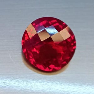 May include: A faceted, round, red gemstone with a brilliant cut. The gem has a deep, rich color and reflects light, creating a sparkling effect. The gemstone is set against a neutral background.