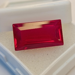 May include: A radiant, rectangular-cut, ruby-red gemstone. The gem is set in a white plastic case with a soft, textured interior. The gemstone's facets reflect light, showcasing its clarity and rich colour.