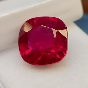 Könnte beinhalten: Ein kissenförmiger, facettierter Rubin-Edelstein. Der Edelstein hat eine leuchtende, tiefrote Farbe mit mehreren Facetten, die das Licht reflektieren. Der Rubin ist vor einem weißen Hintergrund platziert.