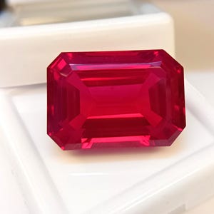 Könnte beinhalten: Ein facettierter, rechteckig geschliffener Rubin-Edelstein. Der Edelstein hat eine leuchtende, tiefrote Farbe und ist vor einem weißen Hintergrund platziert. Die Facetten des Edelsteins reflektieren das Licht.