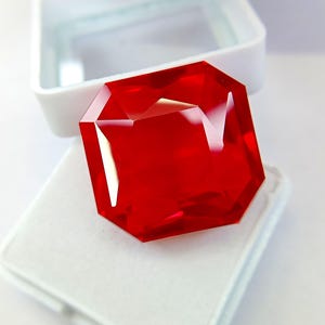 Puede incluir: Una gran gema roja rectangular facetada. La gema se muestra en una caja blanca, mostrando su talla brillante y su color vibrante. Las facetas de la gema reflejan la luz, creando un efecto brillante.