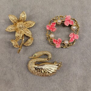 Peut inclure: Trois broches vintage : une fleur dorée, une couronne de fleurs roses et de fausses perles, et un cygne avec un œil vert émeraude. Les broches sont en métal et sont probablement des bijoux fantaisie.
