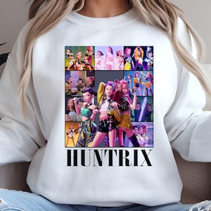 Peut inclure: Sweat-shirt blanc orné d'un collage coloré de personnages animés. Le motif présente plusieurs images de femmes dans diverses poses et tenues, avec le mot "HUNTRIX" en lettres noires grasses en bas.