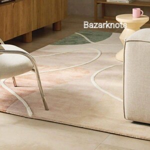 May include: A beige area rug with an abstract design featuring curved white lines and patches of pink and green. The rug is in a room with furniture and the text "Bazarknots" is visible.