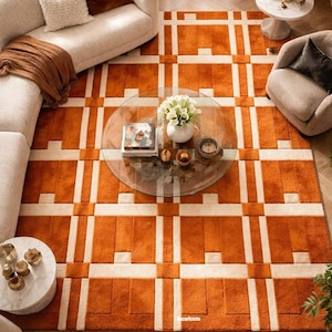 May include: An overhead shot of a living room featuring a large orange and cream geometric patterned rug. The rug is the focal point, complemented by cream-colored sofas, a glass coffee table, and accent chairs. Decorative pillows and a side table with a lamp complete the scene.