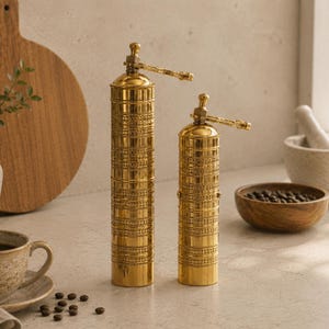 May include: Two tall, cylindrical brass spice grinders with a polished gold finish and intricate horizontal etched patterns. Each grinder features a slender metal handle at the top for manual operation, standing upright on a flat surface.